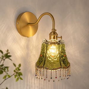 May include: A wall-mounted lamp featuring a brushed gold metal fixture with a curved arm and a bell-shaped, translucent green glass shade. The shade has an embossed floral pattern and is decorated with a fringe of small, multi-colored beads.