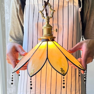 Peut inclure: Une suspension artisanale avec un abat-jour en vitrail aux tons jaune et orange. La lampe est dotée d'une monture en laiton et de perles noires décoratives. L'abat-jour présente un motif floral avec des bordures noires et des accents dorés.