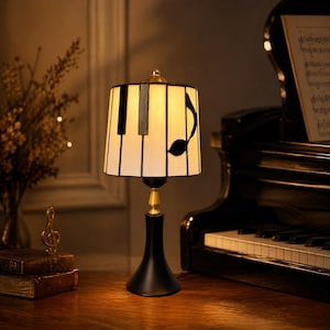 May include: A decorative table lamp with a piano key design on the lampshade and a musical note. The lamp has a black base and a gold accent. A grand piano is in the background.
