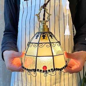 Peut inclure: Suspension décorative avec abat-jour en vitrail. L'abat-jour est de couleur jaune chaud, avec des accents décoratifs noirs et un bijou rouge. Le luminaire a un dessus couleur laiton et un cordon marron.