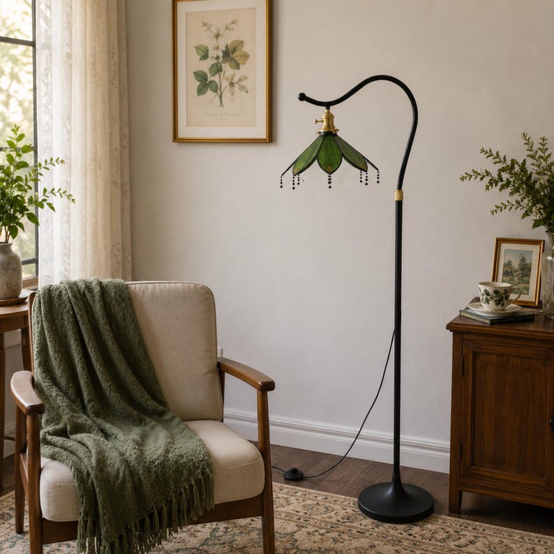 May include: A floor lamp with a black metal stand and a curved arm holding a green stained glass shade shaped like a flower petal. The shade features small black beaded accents hanging from the edges and a brass-colored socket fixture.