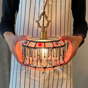 May include: A stained glass pendant light with a pink and white shade, accented with colourful gems and hanging crystal beads. The light fixture has a brass-coloured top and a decorative chain fringe. The light is turned on, illuminating the glass.