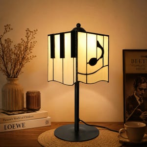 May include: A black piano-themed table lamp with a square shade. The shade features a piano key design and a musical note. The lamp has a black base and stem. The lamp is on a wooden surface.