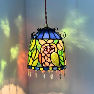 May include: Stained glass pendant lamp featuring an owl design. The lamp has a blue top, a yellow and green body, and a brown cord. Decorative metal accents and dangling leaf charms add to the design. The lamp is approximately 10 inches tall.