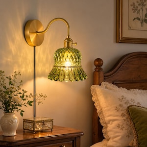 May include: A wall-mounted lamp featuring a brushed brass finish arm and a circular base. The light fixture includes a bell-shaped, textured green glass shade with a decorative beaded trim along the bottom edge, emitting a warm, soft glow.
