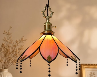 May include: A hanging pendant lamp featuring a petal-shaped stained glass shade in soft pink and orange hues. The fixture includes a brass-toned metal top with decorative filigree accents and dangling black beaded strands along the lower edges.