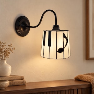 May include: A wall-mounted lamp featuring a curved black metal arm and a cylindrical shade. The shade is constructed from white glass panels arranged to resemble piano keys, with a prominent black musical note graphic on the side.