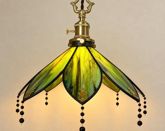 May include: A pendant lamp featuring a stained glass shade with yellow and green marbled panels, framed in dark metal. The lamp includes a brass-toned fixture with a decorative top loop and hanging black beaded accents along the lower edge.
