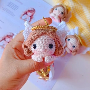 Puede incluir: Un llavero de ángel de crochet con un halo y alas blancas. El ángel tiene el pelo marrón, la cara rosa y ojos grandes marrones. El llavero está unido a un anillo dorado.