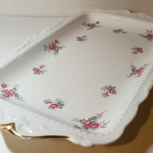 May include: A rectangular white porcelain serving tray with a scalloped edge and gold trim. The tray is decorated with clusters of pink roses and green leaves. The tray measures approximately 30 cm long and 20 cm wide.