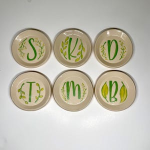 May include: Six small, round, off-white ceramic dishes with green painted letters and botanical designs. Each dish features a different letter: S, K, D, T, M, and B. The letters are surrounded by green leaves and floral accents.