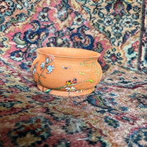 Könnte beinhalten: Ein kleiner Terrakotta-Topf mit handbemaltem Design aus weißen Blumen, grünen Blättern und roten und schwarzen Marienkäfern. Der Topf steht auf einem gemusterten Teppich.