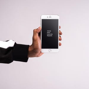 May include: A white smartphone with a black screen displaying the text "Think. Plan. Execute. Repeat." is held in a hand. The person is wearing a black and white sleeve. The background is a solid light grey.