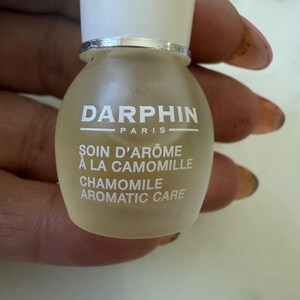 May include: A small, round, frosted glass bottle with a white screw-on cap. The label features the text DARPHIN PARIS, SOIN D'ARÔME À LA CAMOMILLE, and CHAMOMILE AROMATIC CARE printed in white lettering on the front of the bottle.