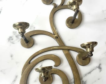 May include: A wall-mounted brass candelabra featuring five circular candle holders attached to a curved, swirling metal frame. The structure has a polished metallic finish with visible screw holes for mounting, showcasing an intricate, flowing design.