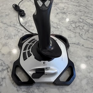 May include: A black and silver Logitech Extreme 3D Pro joystick featuring a central handle with multiple gray buttons, a top-mounted hat switch, a base with a throttle lever, and a USB cable extending from the rear of the device.