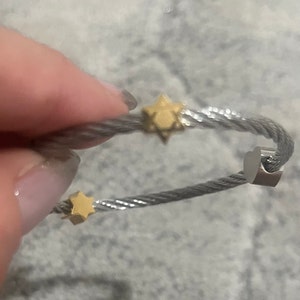 Puede incluir: Un brazalete de cable plateado y dorado con un dije de corazón y dos dijes de estrella.