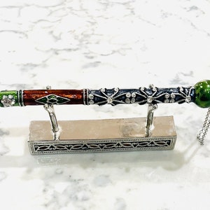 May include: A silver-toned decorative pen stand with a silver-toned pen with a green, brown, and black enamel design. The pen has a hand pointing to the right and a green gemstone at the end.