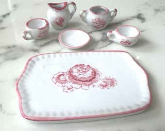 Puede incluir: Un juego de té en miniatura de porcelana que incluye una bandeja rectangular blanca, una lechera, un azucarero, dos tazas pequeñas y un platillo. Cada pieza presenta un estampado floral rosa y un borde pintado en rosa.