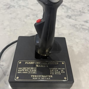 May include: A black Thrustmaster Flight Control System Mark I joystick with a silver top button, a red trigger, and a red thumb button. The base features a metal plate with technical specifications and the text: FLIGHT CONTROL SYSTEM MARK I, THRUSTMASTER, MADE IN THE U.S.A.