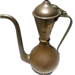 May include: A hammered copper-colored metal pitcher featuring a bulbous base, a tall tapered neck, a long curved spout, and a hinged lid with a small knob handle. The vessel has a metallic finish with visible texture and a curved side handle.