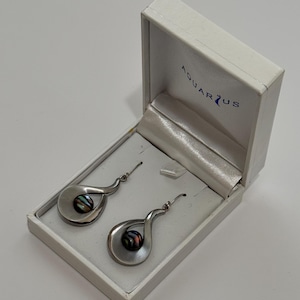 Peut inclure: Une paire de boucles d'oreilles pendantes argentées avec une monture en forme de goutte et une perle centrale sombre et irisée. Les boucles d'oreilles sont présentées dans un écrin blanc avec le nom de marque AQUARIUS imprimé en bleu à l'intérieur.