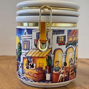 May include: A white ceramic canister with a gold-toned metal latch and lid. The exterior features a colourful painted scene of a Mediterranean-style street cafe with people sitting at tables, a server behind a counter, and white buildings.