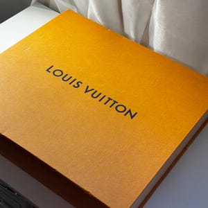 以下が含まれることがあります： 濃い青色で「LOUIS VUITTON」と書かれた長方形のオレンジ色の箱。白い表面に置かれ、背景には白い布があります。