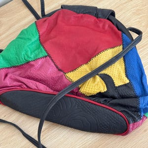 Puede incluir: Una mochila de patchwork multicolor con paneles de cuero texturizado en rojo, verde, amarillo, azul, rosa y negro. El bolso incluye una base de cuero negro con un patrón grabado sutil, una solapa superior y correas negras delgadas.