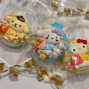 May include: Three character-themed decorations in clear plastic packaging. The characters include a yellow dog with a brown hat, a white bunny with a blue bow, and Hello Kitty with a red bow. Gold leaf garland decorates the background.