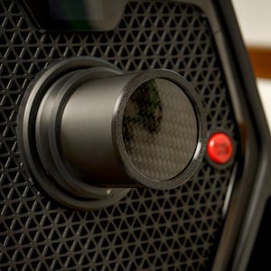 Può includere: Primo piano di un dispositivo nero con un motivo geometrico e una lente cilindrica. Un pulsante rosso è visibile sul lato. La parola "PRO" è stampata nella parte inferiore del dispositivo.