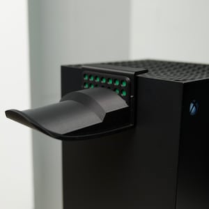 May include: A black gaming console with a green vent and a black plastic cover over the vent.