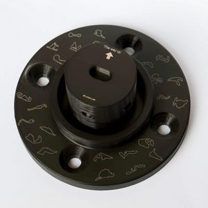 May include: Black circular base with a smaller black cylinder on top. The base has a ring of small race track outlines around the edge. The cylinder has the text "This way up" and an arrow pointing upwards. The cylinder also has the text "PLAYER LUXE" on the side.