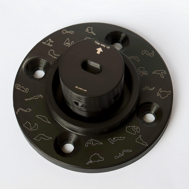 May include: Black circular base with a smaller black cylinder on top. The base has a ring of small race track outlines around the edge. The cylinder has the text "This way up" and an arrow pointing upwards. The cylinder also has the text "PLAYER LUXE" on the side.