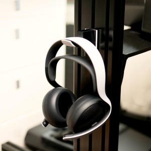 以下が含まれることがあります： 白と黒のヘッドセットが、黒い金属製のスタンドに掛けられています。ヘッドバンドは白、イヤーカップは黒です。