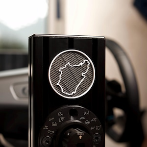 Puede incluir: Un dispositivo de metal negro con un emblema circular plateado que presenta un mapa del circuito de carreras de Nürburgring. El emblema está rodeado por el texto "Nurburgring" repetido varias veces. El dispositivo tiene una perilla con una serie de pequeños contornos de pistas a su alrededor.
