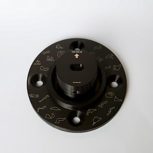 May include: Black metal circular base with a smaller black metal cylinder in the center. The base has a raised edge and is decorated with small outlines of race tracks. The cylinder has a small hole in the center and the text "This way up" above the hole. The text "FLUXLINE" is printed on the cylinder.