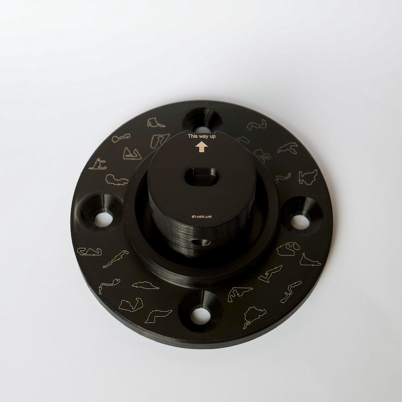 May include: Black metal circular base with a smaller black metal cylinder in the center. The base has a raised edge and is decorated with small outlines of race tracks. The cylinder has a small hole in the center and the text "This way up" above the hole. The text "FLUXLINE" is printed on the cylinder.