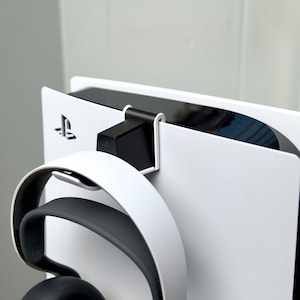 May include: A white PlayStation 5 console with a black headset holder attached to the top. The headset is hanging on the holder. The PlayStation logo is visible on the console.