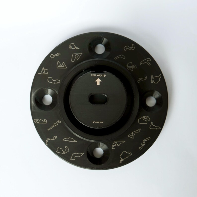 May include: Black circular metal object with a black center and a silver arrow pointing upwards with the text "This way up". The outer edge of the object is decorated with small outlines of race tracks.