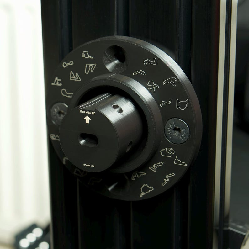 May include: A black metal knob with a circular design featuring outlines of famous race tracks. The knob has a small arrow pointing up with the text "This way up".