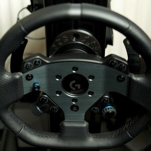 May include: A black racing steering wheel with a silver center plate featuring a G logo. The wheel has multiple buttons and a knob on the left side. The wheel is attached to a black base with a circular dial with various symbols.