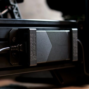 May include: A black power adapter with a textured surface and a series of small, etched symbols. The adapter is attached to a black device with a power cord.