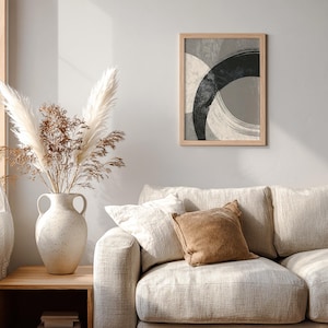 Pode incluir: Uma impressão de arte abstrata emoldurada com formas pretas, cinzentas e bege sobrepostas. A obra de arte é exibida acima de um sofá bege com almofadas decorativas. Um vaso com flores secas está em uma mesa lateral de madeira.