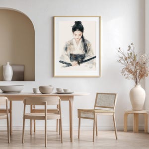 May include: A framed watercolor print depicts a person in traditional Japanese attire holding a katana sword. The artwork features soft, muted colors and is displayed in a gold frame. The image is set in a minimalist interior with a dining table and chairs.