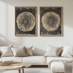 May include: Two framed wall art pieces featuring tree ring designs. The artwork has a dark background with a circular, light brown tree ring pattern. The frames are a light brown color. The art is displayed above a white couch with neutral-colored pillows.