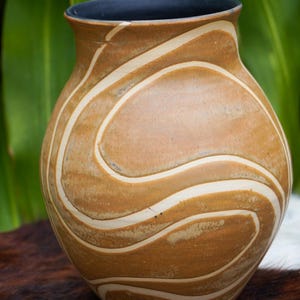 Puede incluir: Un jarrón de cerámica marrón con un esmalte blanco, que presenta un diseño ondulado abstracto. El jarrón tiene un cuerpo redondeado y una abertura ligeramente acampanada.