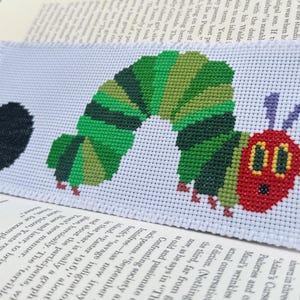 May include: A cross-stitch pattern of a green caterpillar with a red head and black eyes. The caterpillar is curled into a circle shape. The pattern is on white fabric and includes a black circle.