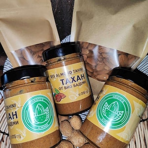 May include: Three glass jars of almond tahini, labeled "Tahini" and "Bio Almond Tahini", with the text "Tahini" and "Bio Almond Tahini" in English and Bulgarian. The jars are filled with a light brown, creamy spread. The jars are sitting on a woven basket filled with almonds.