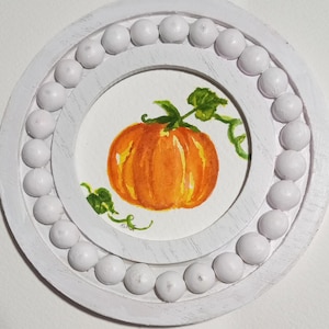 May include: A round, white wooden frame with a watercolor painting of an orange pumpkin with green vines and leaves. The frame has a beaded outer edge. The pumpkin is centered on a white background.